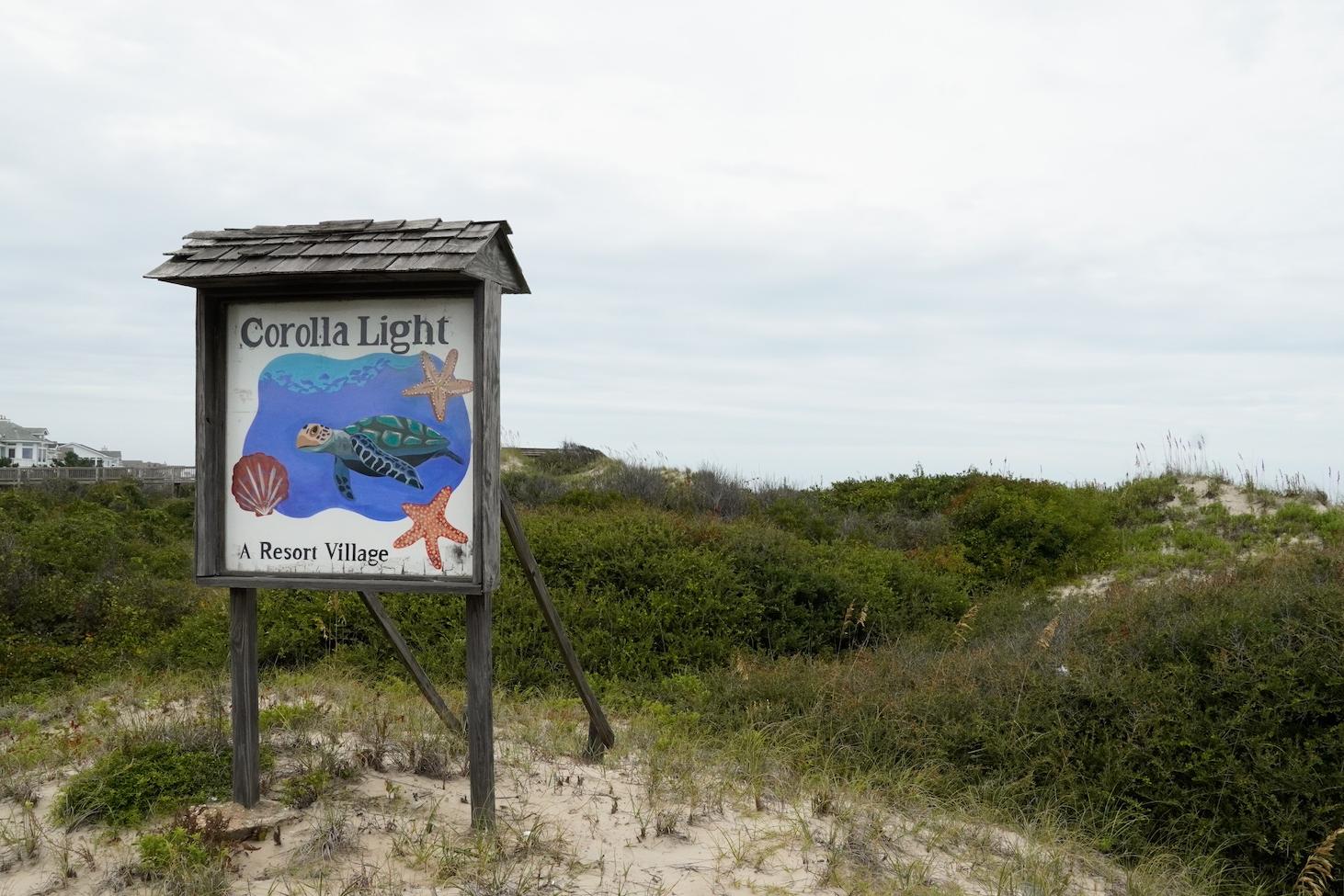 A hand-painted Corolla Light Resort sign on the plant-covered sand dunes between the resort and the Atlantic Ocean.