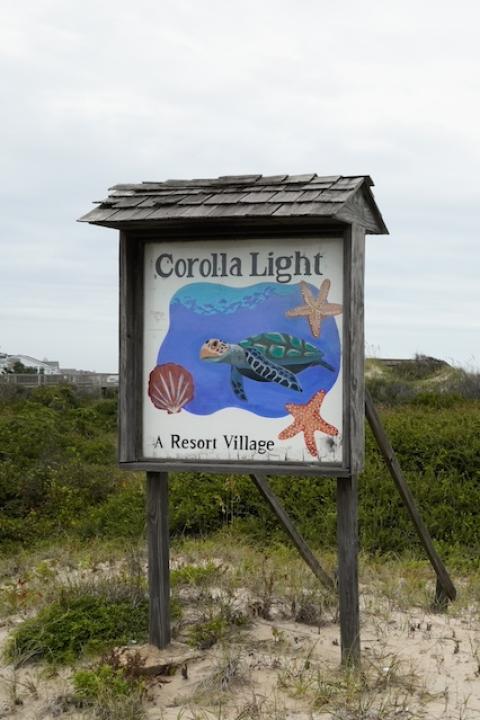 A hand-painted Corolla Light Resort sign on the plant-covered sand dunes between the resort and the Atlantic Ocean.