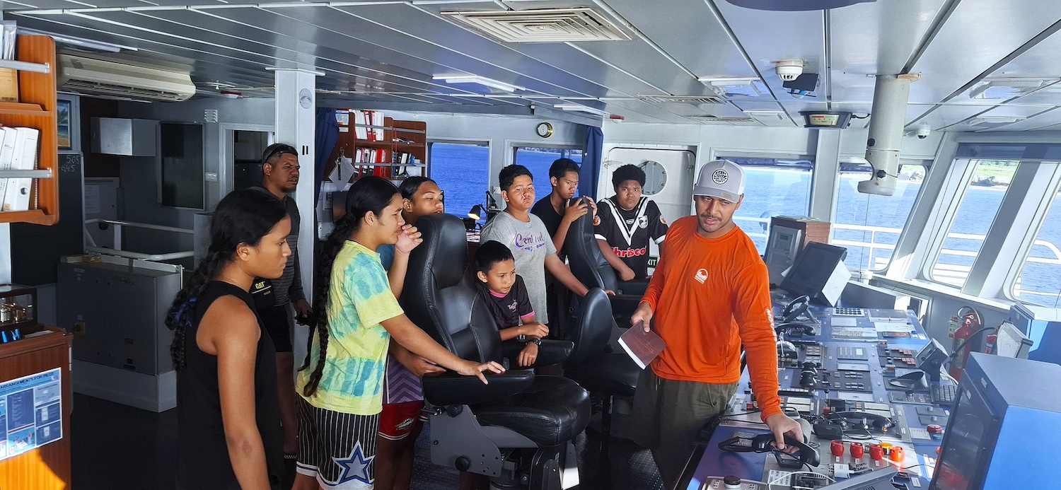 A group of Mauke locals stand in the bridge of a Moana Minerals deep-sea mining vessel looking at the ship's forward controls.