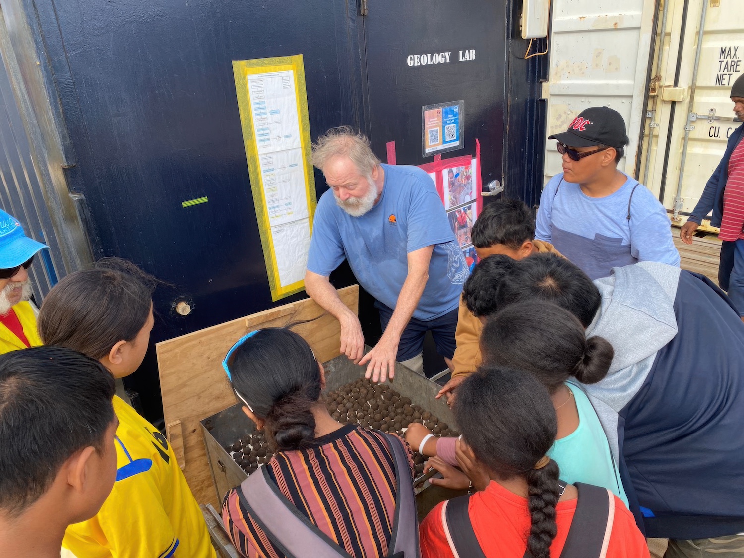 The polymetallic nodules that can be found on the ocean floor, resembling small brown spheres of rock, are displayed in a box while John McIntyre talks about them with Cook Islanders — deep-sea mining