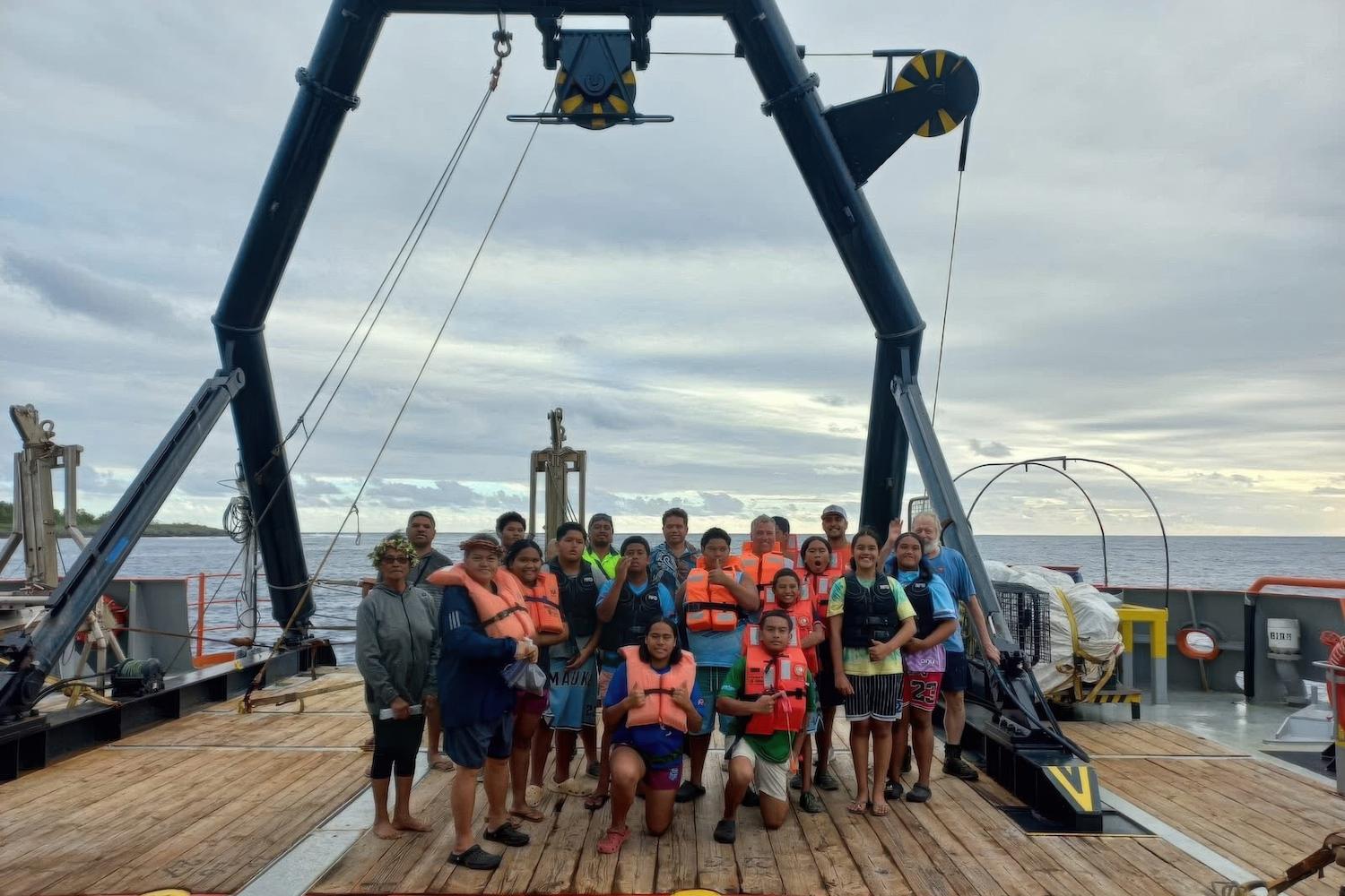 Residents of Mauke, Cook Islands, stand together on the deck of a deep-sea mining boat under a pulley system.
