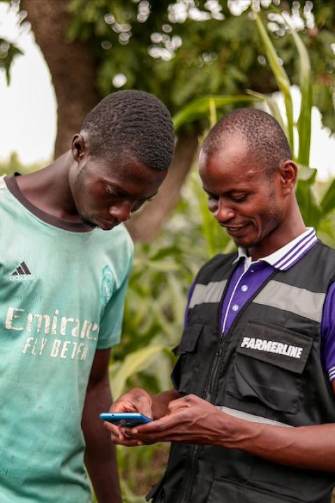 A Farmerline team member and a farmer look down at a smartphone near an agricultural field.