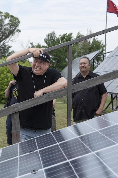 Cody Two Bears smiles while leaning on a solar panel array — energy sovereignty